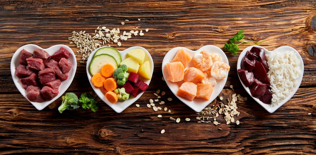 Healthy fresh ingredients for pet food in individual heart-shaped bowls viewed from overhead with chopped raw beef, liver and chicken , mixed vegetables and rains on rustic wood